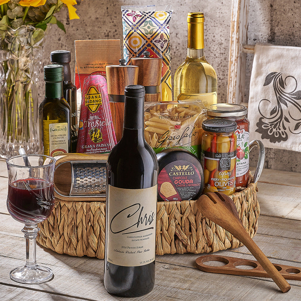 Wine bottle, glass of red wine, and assorted food items in a gift basket on a wooden surface.